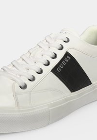 Baskets basses blanches avec lacets blancs, panneau latéral texturé noir et le nom de la marque "GUESS" imprimé en blanc sur le côté.