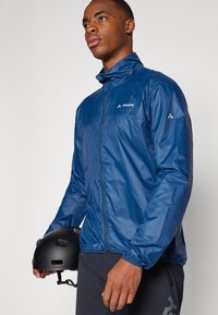 Navy blue lightweight jacket with a high collar, zip front, and elastic cuffs. Features a logo on the chest and a matte finish.