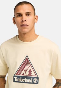 Krémové bavlněné tričko s trojúhelníkovou grafikou obsahující nápis "Rugged Active Gear", designy stromu a hory v bordó a černé barvě.