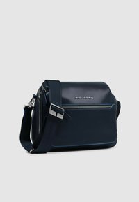 Bolso mensajero de cuero azul marino con un diseño estructurado, herrajes plateados, correa ajustable y un bolsillo frontal con cremallera y costuras azules a contraste.