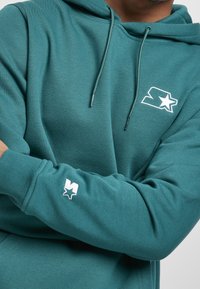 Sudadera con capucha color azul verdoso hecha de tela suave, con un logo en el frente y un pequeño detalle en la manga, con cordones ajustables y puños acanalados.