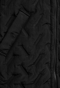 Zwart quilted stof met diagonale gestructureerde strepen, een zichtbare rits aan de rechterkant en een gestikt gewatteerd vak aan de linkerkant.