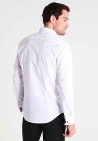 Camicia bianca a maniche lunghe con orlo curvo, caratterizzata da un colletto classico e dettagli con bottoni sui polsini. Tessuto morbido, vestibilità su misura.