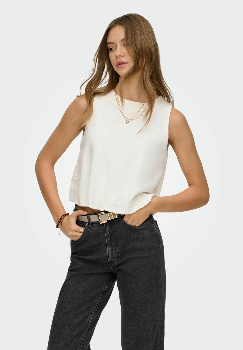 Jeune femme aux longs cheveux portant un haut blanc sans manches, un jean noir taille haute, un collier en or, un bracelet et une ceinture, les mains dans les poches.