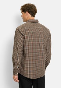 Mann von hinten, der ein braun-, schwarz- und weißkariertes Langarmhemd und schwarze Hosen vor einem schlichten weißen Hintergrund trägt.