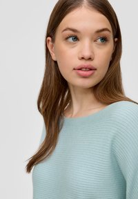 Hellblau gestreifter Pullover mit weitem Ausschnitt. Modell mit langem, glattem braunem Haar in schräger Position, wegschauend. Weiche Textur.