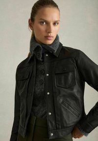 Schwarze Lederjacke mit Druckknopfverschluss, zwei Fronttaschen und einem flauschigen grauen Pullover darunter, der einen hohen Kragen und Knöpfe hat.