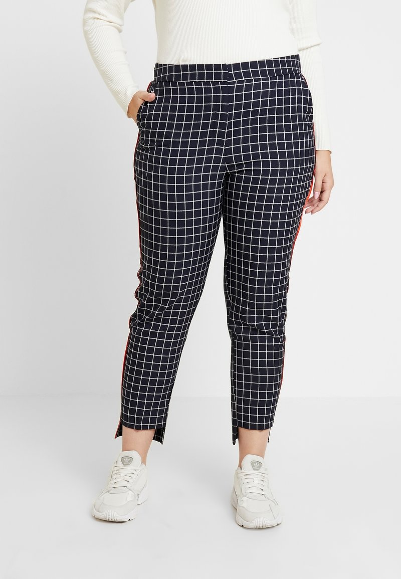 Femme portant un pantalon court à motifs à carreaux noirs et blancs avec des bandes rouges sur les côtés, des baskets blanches, et un pull côtelé blanc.