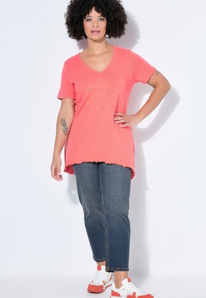 Vrouw met krullend haar die een koraalkleurig T-shirt met V-hals draagt, cropped blauwe jeans, witte en koraal gekleurde sneakers, staand met hand op heup tegen witte achtergrond.