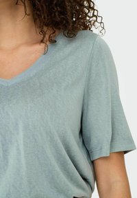 Camiseta verde claro de pico, hecha de una tela suave y texturizada, con mangas cortas y un corte relajado. Costuras visibles a lo largo del escote.