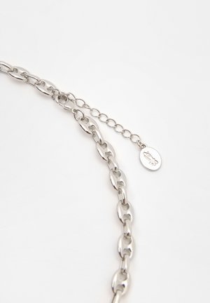 Silveren kædelys i kædeform med ovale led, et lille rundt mærke med logo og justerbar låsemekanisme. Glat metaloverflade.