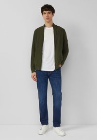 Olijfgroene jas met rits over een wit T-shirt, gecombineerd met blauwe jeans en witte sneakers. Eenvoudig ontwerp, gladde textuur en aansluitende pasvorm.