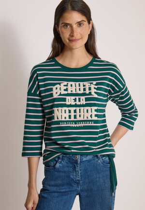 Femme aux longs cheveux bruns portant un t-shirt rayé vert et rose avec un texte en français et un jean bleu, souriante, une main sur la hanche.