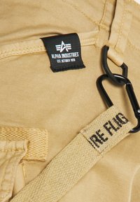 Bežová tkanina s čiernou nálepkou Alpha Industries, čierne kovové klipsy a čiastočne viditeľný béžový tkaný popruh s textom "FIRE FLAG".