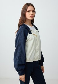 Lichte, twee-kleurige windbreaker jas met een marineblauwe capuchon en mouwen, een crème-kleurig lichaam, elastische manchetten en ritssluiting.