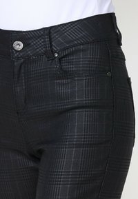 Gros plan d'un pantalon ajusté à carreaux noirs et gris avec bouton, fermeture éclair et détails de poche avant sur une personne portant un haut blanc.