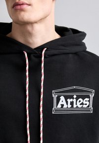 Zwarte hoodie met een wit "Aries" logo met architectonische details. Het heeft gestreepte trekkoorden in rood en beige. Zacht, katoenen materiaal.