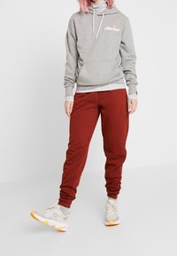 Felpa grigia con cappuccio e logo bianco, abbinata a pantaloni sportivi rossi. L'outfit ha un aspetto sportivo e include sneakers di colore chiaro.