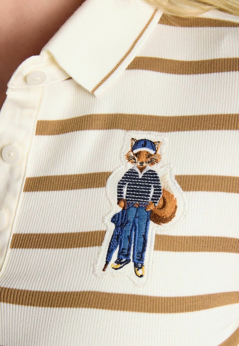 Smeđa i bijela prugasta majica s malim izvezenim lisicom koja nosi kapu, prugastu majicu, plave hlače i drži kišobran.