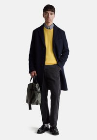 Tmavě modrý vlněný kabát, žlutý svetr, šedé kalhoty, černé boty, držená zelená taška. Kostkovaná košile pod svetrem. Hladké textury.