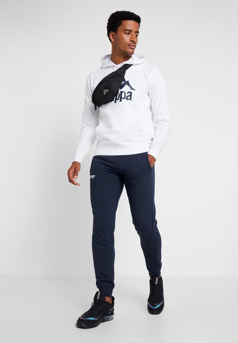 Sudadera blanca con logo en marino, pantalones de chándal en marino y zapatillas negras. Lleva un bolso bandolera negro cruzado sobre el pecho. El modelo está de pie contra un fondo liso.