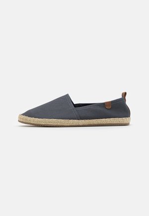 Sive platnene slip-on čevlje z juto vrvjo, ki imajo rjav usnjen detajl na peti in minimalističen dizajn.