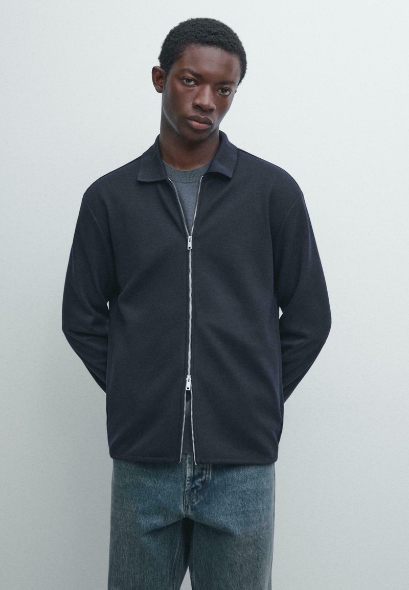 Mørk lynlåstrøje med krave, lavet af blødt stof. Har et lige snit og lange ærmer, kombineret med lyseblå jeans.