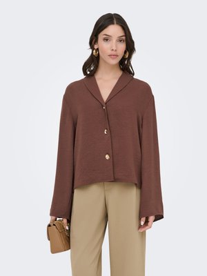 Femme portant un haut marron à manches longues boutonné, pantalon beige, boucles d’oreilles créoles dorées, tenant un petit sac à main beige avec une bandoulière chaîne.