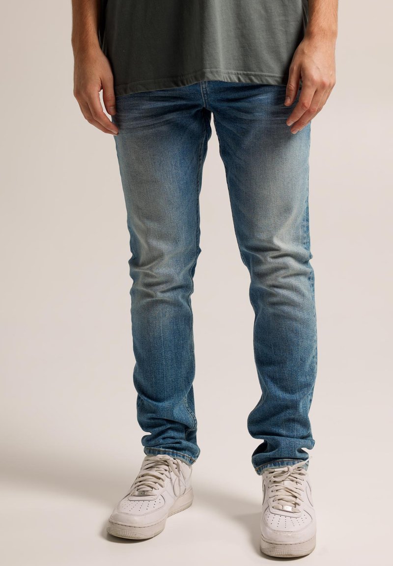 Blaue Denim-Jeans mit schmaler Passform, ausgestattet mit einem verblassten Wascheffekt, subtiler Textur und gebrauchten Look. Kombiniert mit weißen Sneakern.