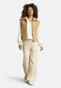 Tan wool blend sleeveless vest over a white long-sleeve shirt, paired with cream-colored wide-leg trousers and white sneakers.