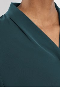 Blusa di un verde scuro con colletto a scialle, texture di tessuto liscia e un design leggermente drappeggiato che mette in risalto linee pulite.