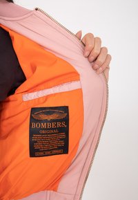 Różowa kurtka bomberka z pomarańczową podszewką. Posiada złoty zamek błyskawiczny oraz czarną metkę z białym napisem wewnątrz kieszeni. Gładka faktura materiału.