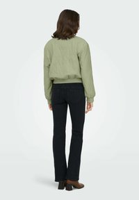 Veste bomber verte avec une finition texturée, associée à un jean évasé bleu foncé, des détails de coutures visibles, et des bottines marron à talons blocs.