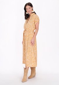 Donna con capelli scuri che indossa un vestito midi giallo a fantasia paisley, stivali da cowboy beige e grandi orecchini tondi, in piedi contro uno sfondo bianco.