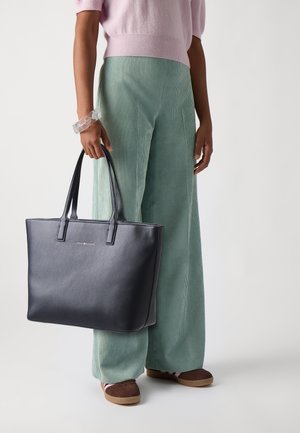 Sac fourre-tout en cuir marine avec deux poignées, forme rectangulaire, texture lisse. Le modèle porte un pantalon en velours côtelé vert menthe et des chaussures marron.