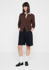 Brun kabelstrikket cardigan over en lys stripete skjorte, sammen med marineblå shorts. Sorte loafers og hvite sokker fullfører antrekket.