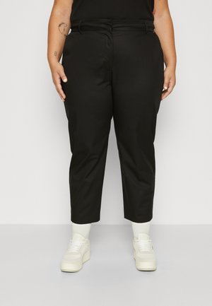 Person trägt schwarze, hoch taillierte, gekürzte Hose, weiße Socken und weiße Plateau-Sneaker und steht vor einem schlichten hellen Hintergrund.