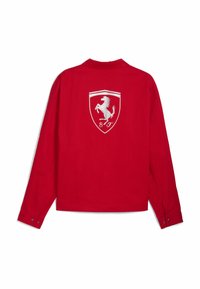 Cămășă roșie cu mâneci lungi, având un logo alb Ferrari și un model cu scut pe spate. Fabricată dintr-un material neted, cu manșete cu nasturi.
