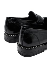 Mocasines de charol negro con borde con tachuelas alrededor de la suela y tacones bajos de apilamiento, mostrados desde la parte posterior sobre un fondo blanco.