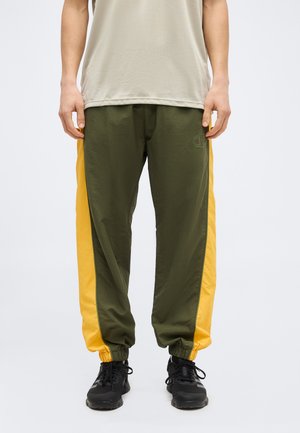 Man wearing olive green and yellow track pants, beige shirt, and black athletic shoes, standing against plain background.