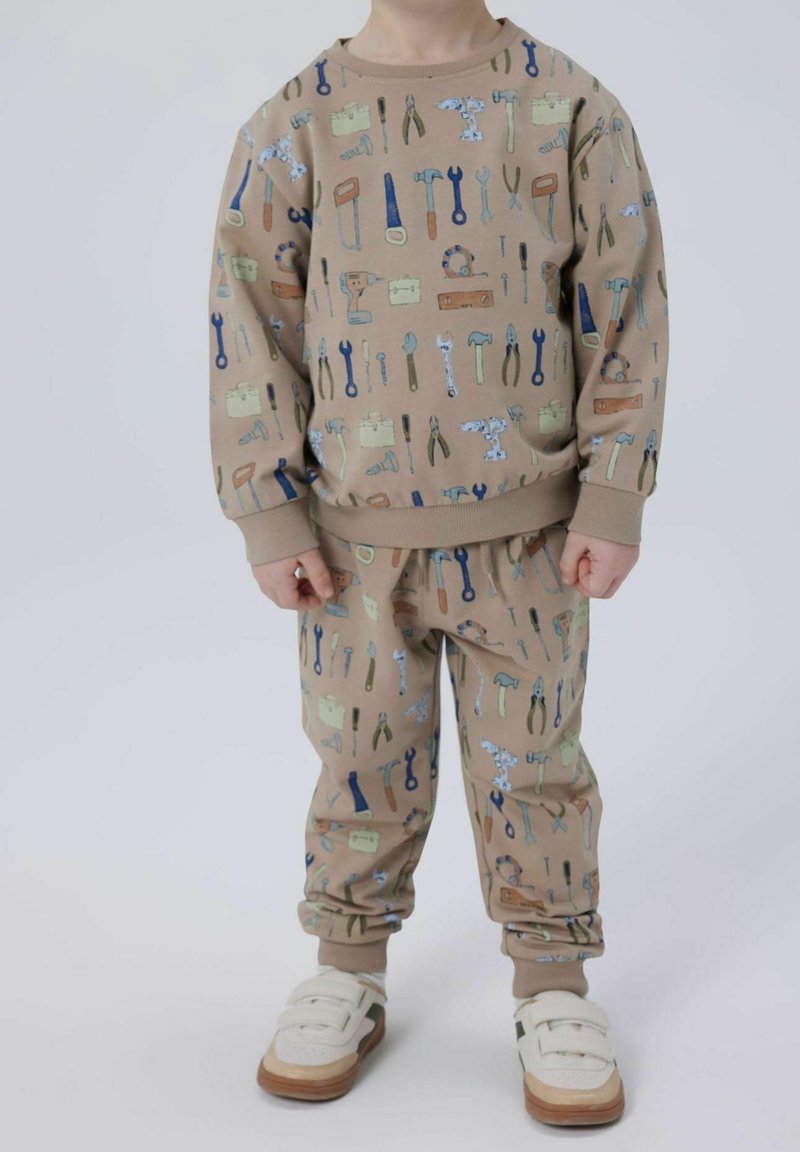 Enfant portant un sweat-shirt beige assorti et un pantalon avec des imprimés colorés d'outils de construction, et des baskets blanches à scratch.