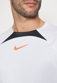 Koszulka sportowa Nike w kolorze białym z czarną wstawką przy dekolcie oraz pomarańczowym logo swoosh na piersi.