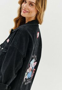 Czarna dżinsowa kurtka z dużą graficzną aplikacją kwiatową z tyłu, w pastelowych kolorach i z napisem, wykończona akcentami w czerwonym kolorze.
