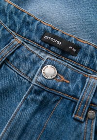 Blå denimjeans med orange sömmar, märkta "ombre" på en svart etikett, utrustade med en metallknapp med gravyrer och en rak design.