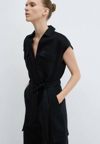 Zwarte linnen jumpsuit met korte mouwen, twee borstzakken, een gestrikt tailleband en een wijd-pijp ontwerp. Gladde textuur, minimalistische stijl.