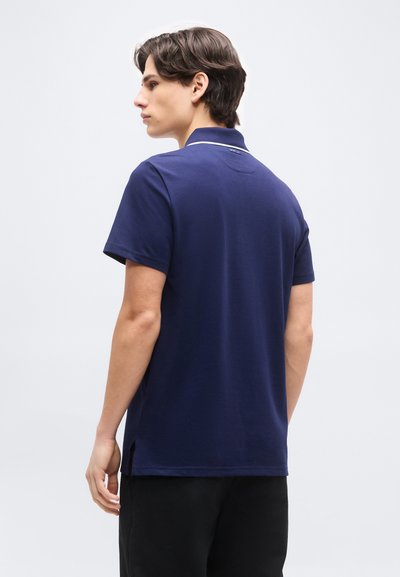 Νεαρός άνδρας με την πλάτη, ντυμένος με μπλε σκούρο κοντομάνικο πόλο και μαύρο παντελόνι, μπροστά σε απλό ανοιχτό φόντο.