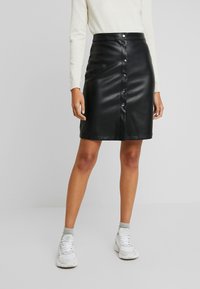 Jupon en simili cuir noir avec une taille haute, fermeture à bouton à l'avant et texture lisse. Associé à un pull léger et des baskets blanches.