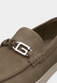 Καφέ μοκασίνι από σουέντ με πλεγμένη λεπτομέρεια δέρματος και ασημί μεταλλικό διακοσμητικό «G» στην κορυφή.