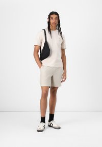 Tricou beige cu mânecă scurtă și grafică cu palmier, pantaloni scurți gri deschis cu dungi, geantă de umăr neagră și adidași albi cu accente gri și negre.