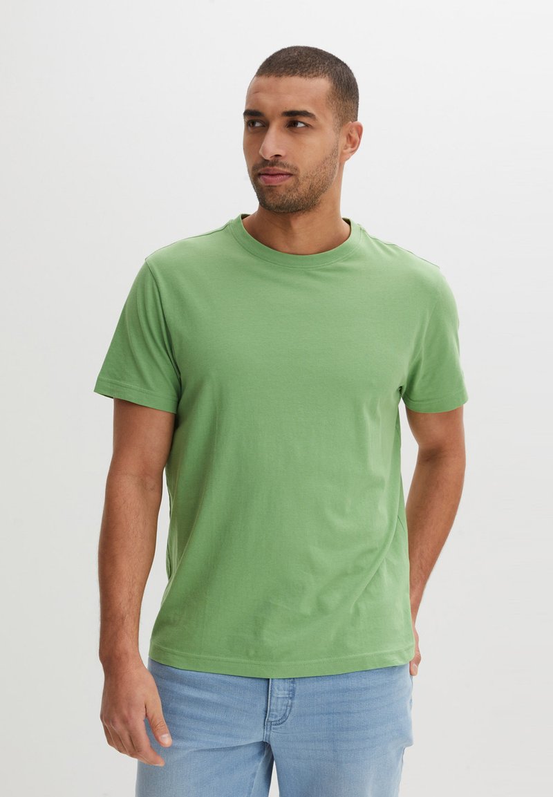 T-shirt verde in cotone con scollatura a girocollo, maniche corte e vestibilità rilassata. Texture liscia, cuciture minime, abbinato a jeans azzurri chiari.